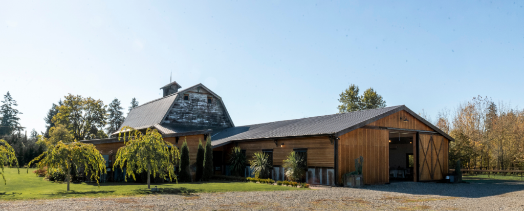 A large, wood-sided event barn with a metal roof and open sliding doors, situated on a grassy lawn with a gravel driveway.